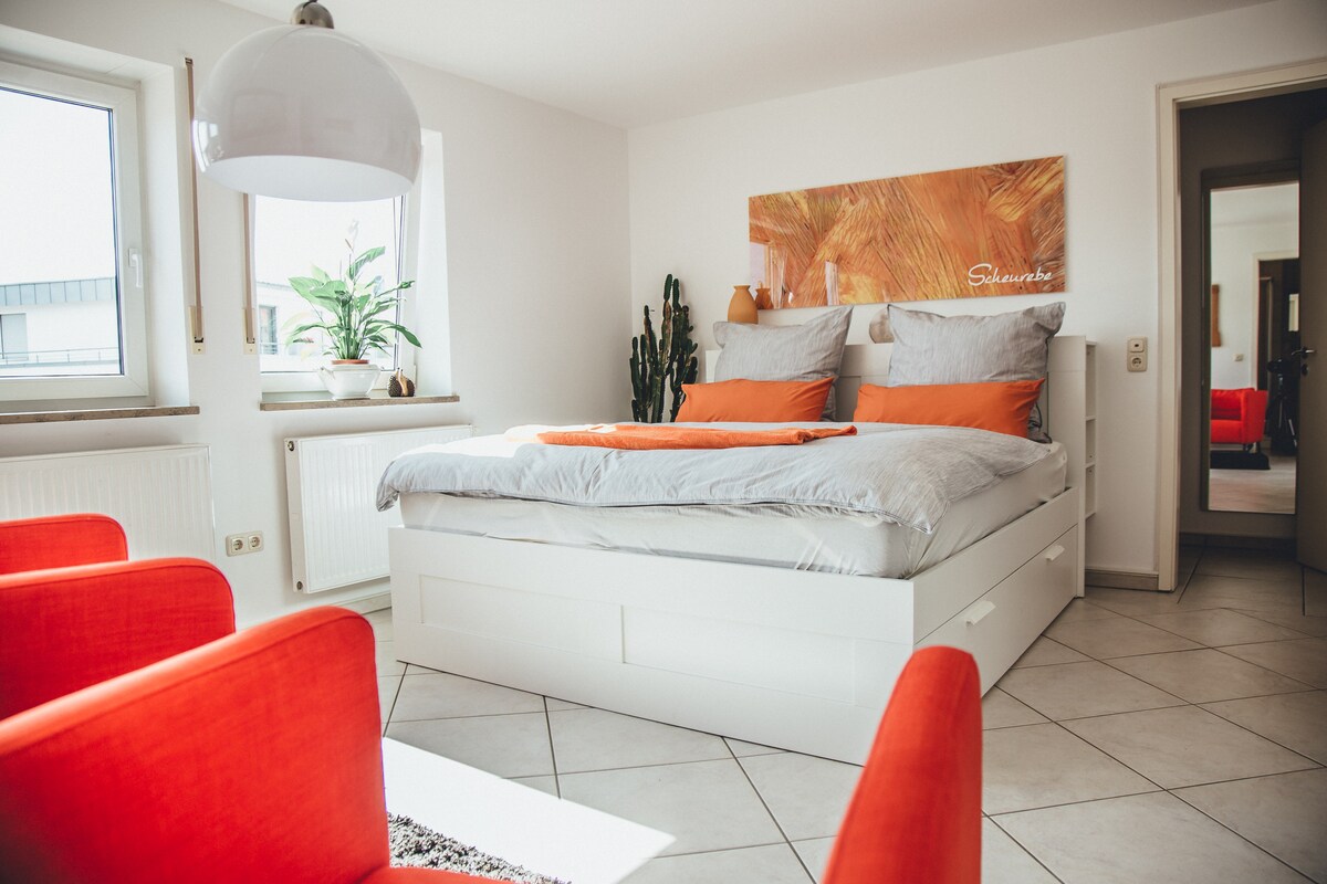 A light and airy bedroom features a spacious bed with gray bedding and decorative orange pillows. Two large windows allow natural light to fill the room, complemented by a leafy plant. A mirrored closet and red chairs add to the welcoming atmosphere.