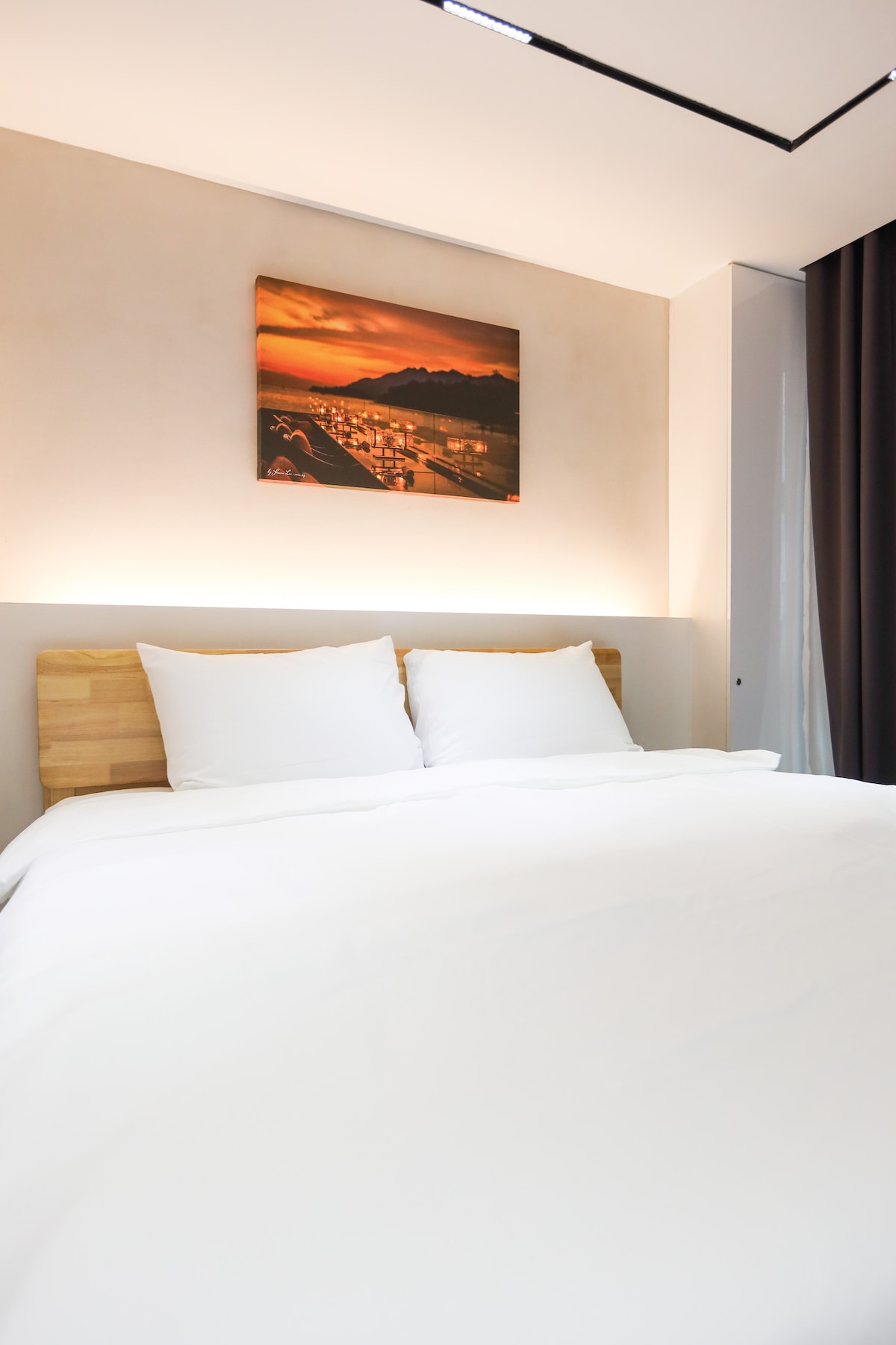 A welcoming bedroom features a king size bed adorned with a crisp white comforter and two plush pillows. A soft ambient light emanates from behind the headboard, complemented by a serene artwork depicting a sunset. Dark curtains frame the window, adding a touch of sophistication.
