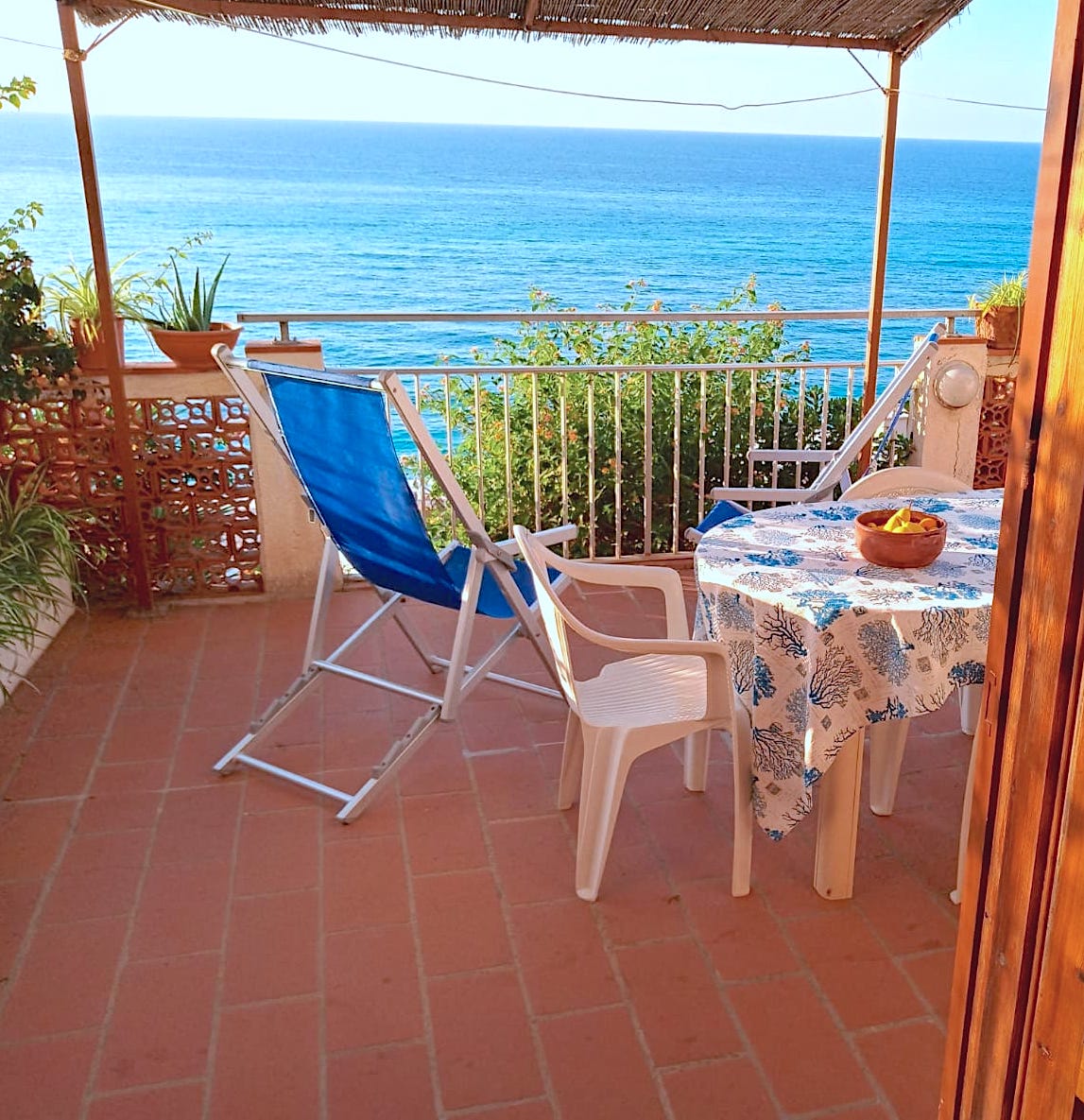 A terrace is seen overlooking the sea, featuring a blue reclining chair and a white table with a floral tablecloth. A bowl of fruit is placed at the center of the table. Greenery and the blue ocean create a serene backdrop under the shade of a woven canopy.