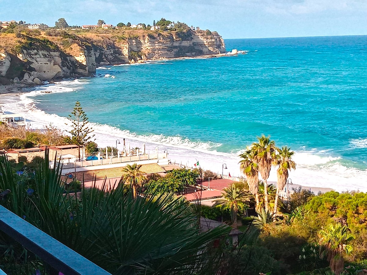 A scenic view of the coastline is presented, featuring the vibrant blue sea meeting sandy shores. Lush greenery and palm trees are visible in the foreground, while gentle waves create white foam along the beach. The cliffside adds depth to the natural landscape.