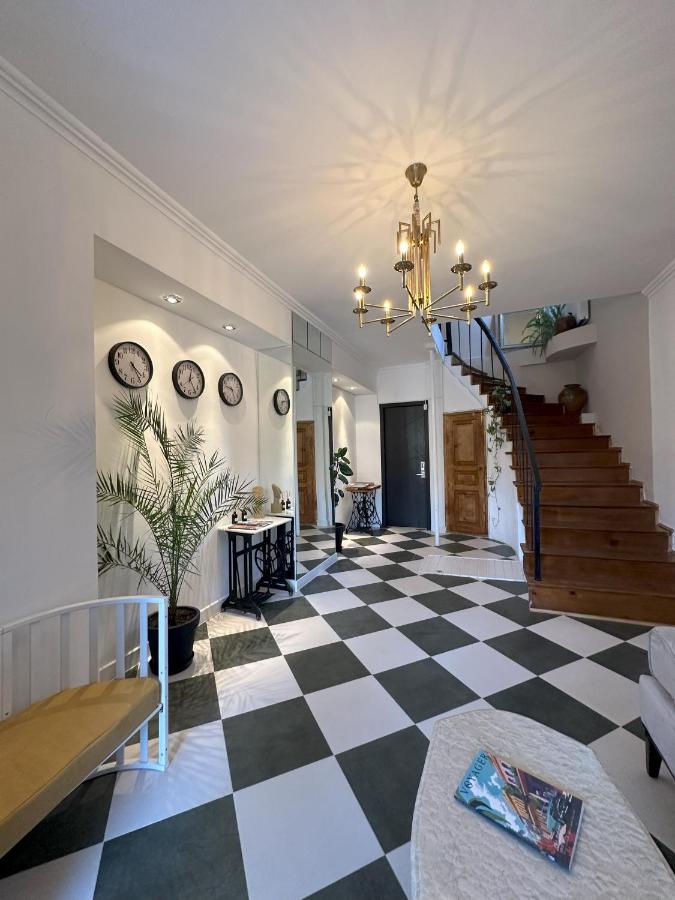 A spacious hallway is presented, featuring a checkerboard floor with alternating dark and light tiles. A staircase with wooden steps leads to the upper level. Plants and decorative elements add a touch of greenery, while wall-mounted clocks and light fixtures contribute to the inviting atmosphere.