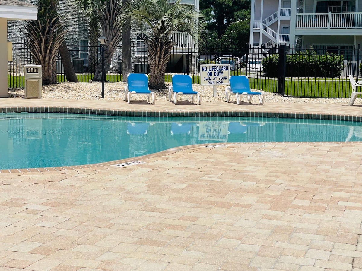 A spacious pool area features a clear blue swimming pool surrounded by paved decking. Several lounge chairs with blue cushions are positioned beside the water, while palm trees provide a natural backdrop. A safety sign indicating no lifeguard on duty is visible.