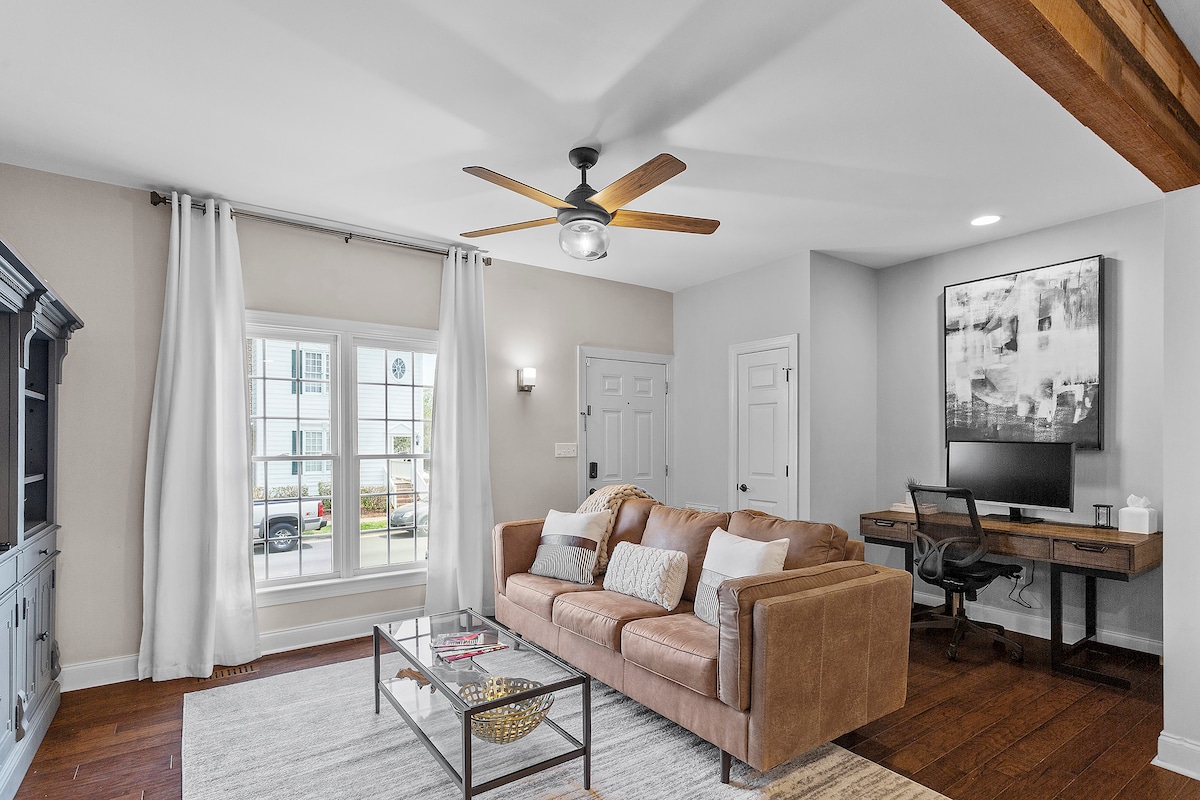 A comfortable living area features a brown leather sofa with decorative pillows, paired with a glass coffee table. A ceiling fan enhances airflow above, while natural light filters through large windows adorned with white curtains. A workspace is positioned nearby with an office desk and chair.