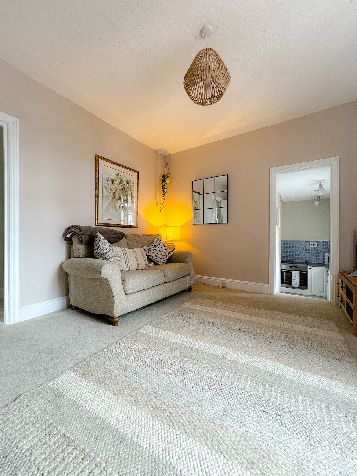 A welcoming lounge area features a comfortable sofa adorned with decorative cushions, a soft rug covering the floor, and a warm glow from a nearby lamp. A wall-mounted mirror enhances the space, with a doorway leading to the kitchen visible in the background.