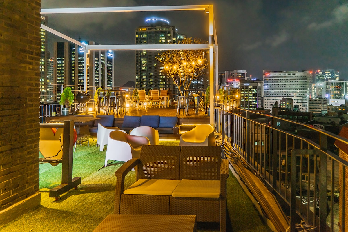 An inviting rooftop space is shown, featuring comfortable seating arrangements and a barbecue area. Soft lighting enhances the atmosphere, while views of the city skyline illuminate the background. A grassy surface adds a touch of greenery to the urban setting.