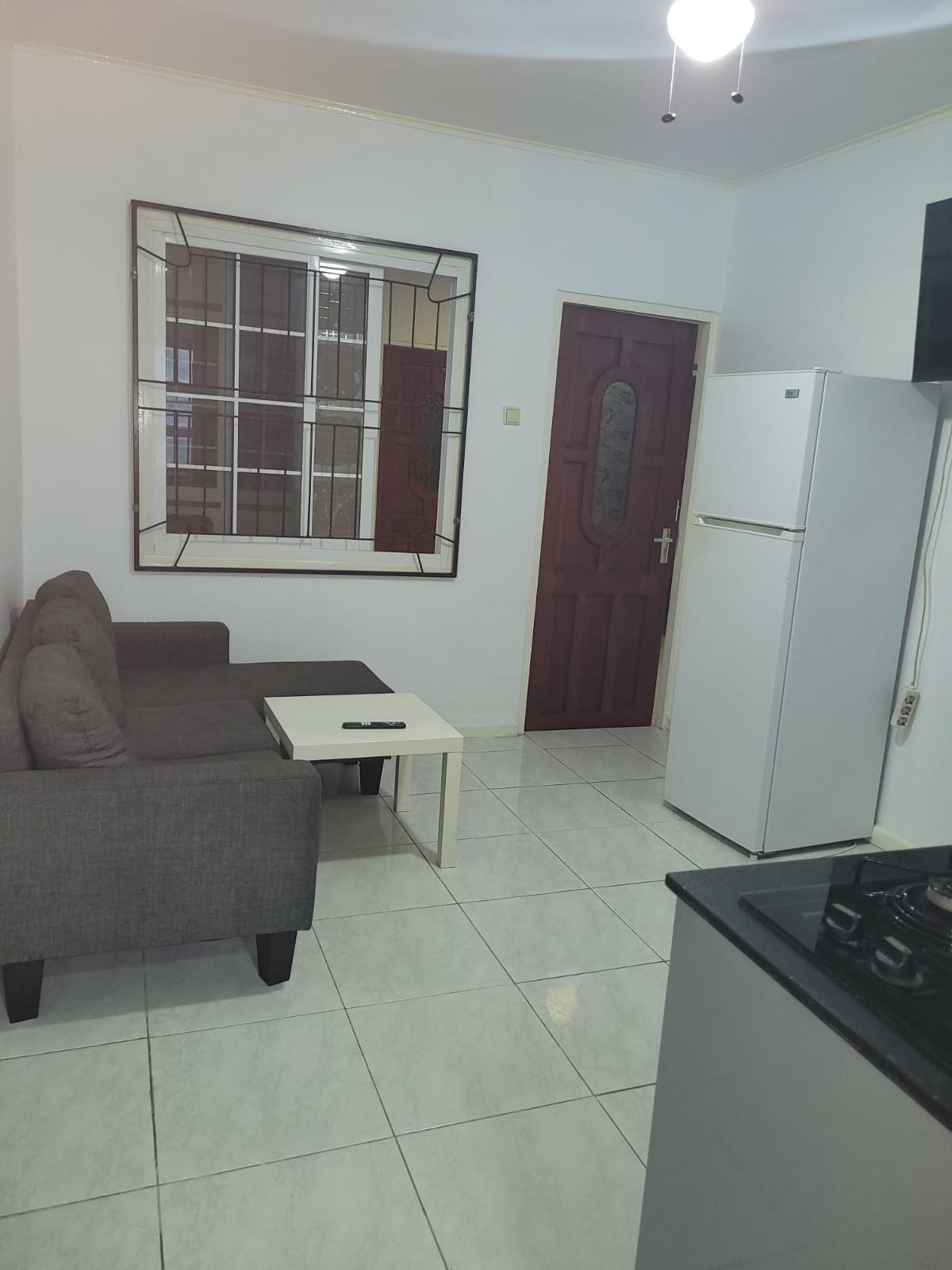 A compact living area is equipped with a gray sofa, small white coffee table, and a door leading to another space. A refrigerator stands against the wall next to a kitchen area featuring a stove. The tiled floor and light walls enhance the room's brightness.