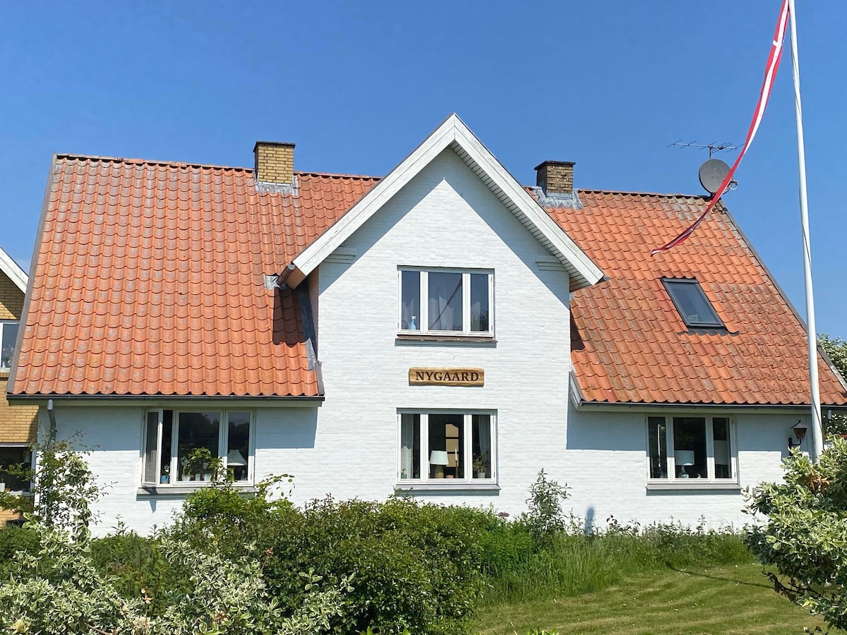 A charming two-story house features white exterior walls with a distinctive red tile roof. Large windows allow natural light to fill the interior. The house is surrounded by well-maintained greenery, emphasizing its inviting presence. A flagpole is visible in the foreground.