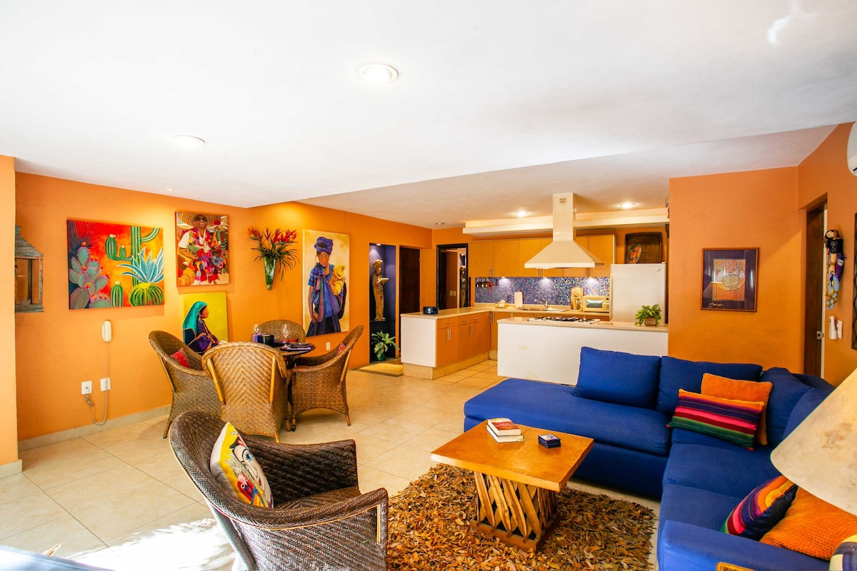 A spacious living area is depicted with a comfortable blue sofa adorned with colorful cushions. A wooden coffee table sits at the center, surrounded by woven chairs. Brightly colored artwork is displayed on the orange walls, complementing the open kitchen in the background.