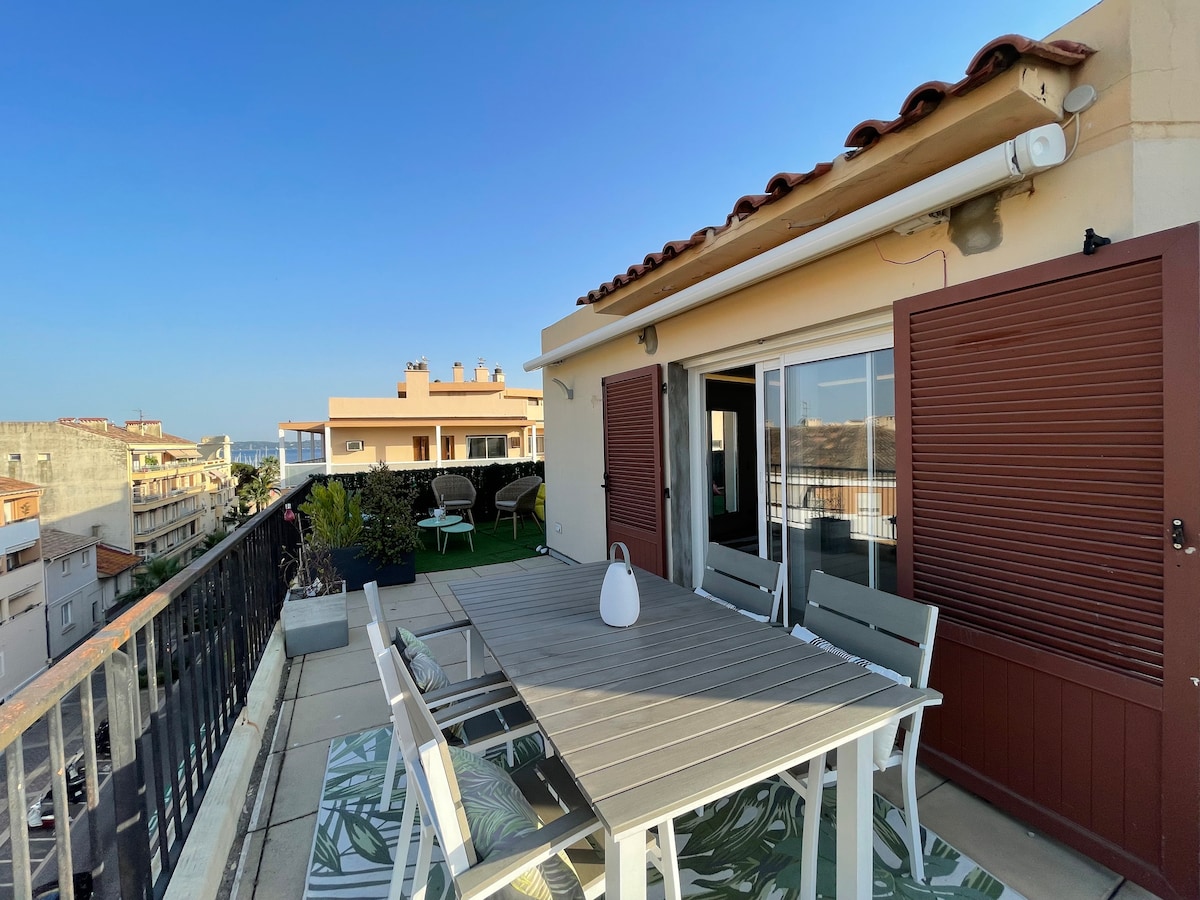 The spacious terrace is highlighted by a large dining table surrounded by several chairs. Potted plants add greenery to the space, with panoramic views of the surrounding buildings and blue sky. Sliding glass doors open to the interior, allowing for seamless indoor-outdoor living.