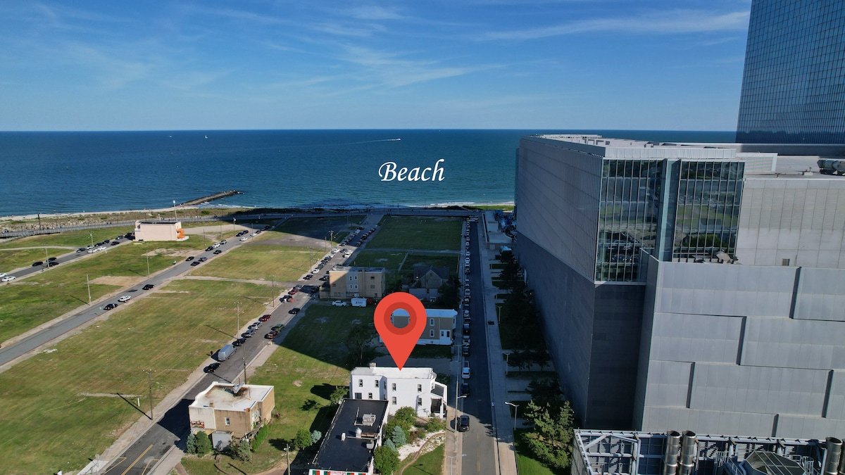 An aerial view captures a white building positioned near the shoreline, with direct access to the beach visible. The expansive blue ocean meets the horizon, while nearby structures and green spaces provide context. A marked location indicates the beach area for reference.