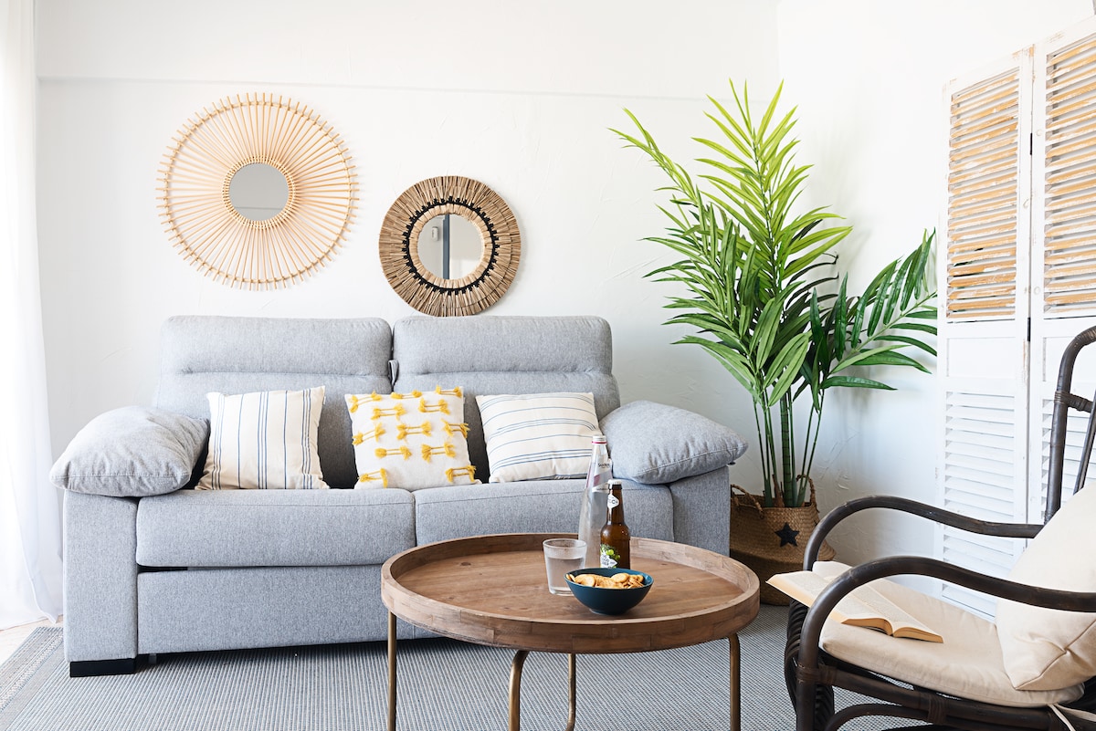 A comfortable living area features a light grey sofa adorned with patterned cushions. Two round mirrors hang on the wall, reflecting natural light. A tall green plant adds a touch of nature, while a round wooden coffee table supports a bowl and beverages.