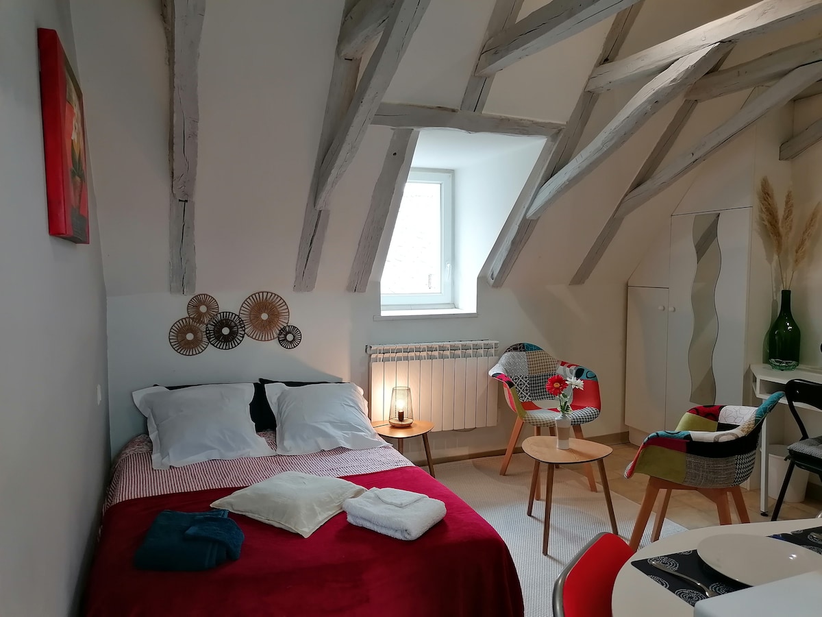 A studio space features exposed wooden beams and a cozy sleeping area with a spacious bed dressed in red and white linens. A small seating arrangement is visible, with two chairs beside a round table. A gentle light is provided by a bedside lamp.
