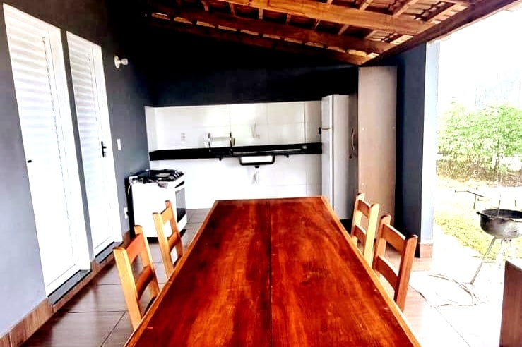 A spacious dining area features a long wooden table surrounded by several wooden chairs. The modern kitchen is partially visible in the background, showcasing white cabinetry and appliances. Natural light filters in through open double doors, highlighting the inviting setting.
