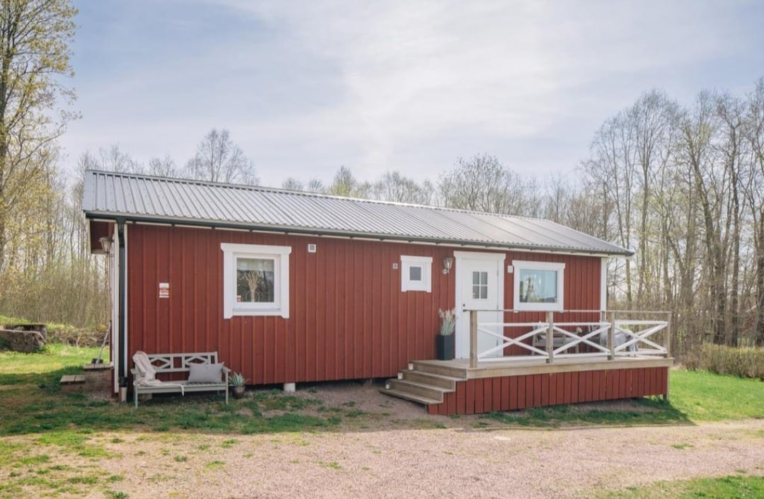 A charming red wooden house with a metal roof is featured. Steps lead up to a welcoming front porch framed by a railing. Green grass surrounds the property, while trees provide a natural backdrop, contributing to a serene countryside environment.
