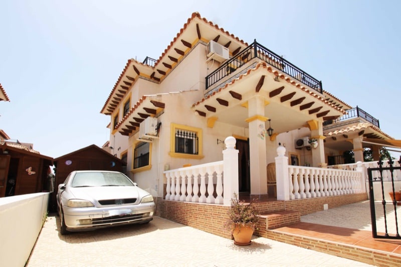 The exterior of the town house is presented, featuring a combination of decorative elements and a welcoming entrance. A car is parked in the driveway, while potted plants add a touch of greenery. The balcony above offers additional outdoor space complemented by terracotta tiles.