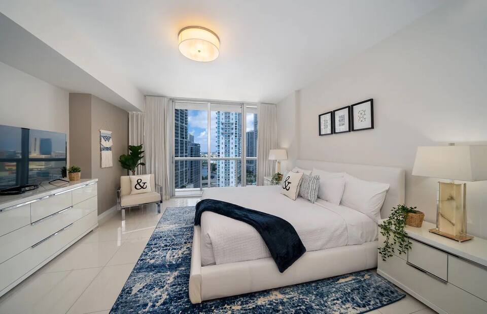 A spacious bedroom is furnished with a large bed, dressed in white linens and decorative pillows. A flat-screen television is mounted on a sleek white dresser. Large windows offer city views, while a soft blue and white rug adds warmth to the polished floor.