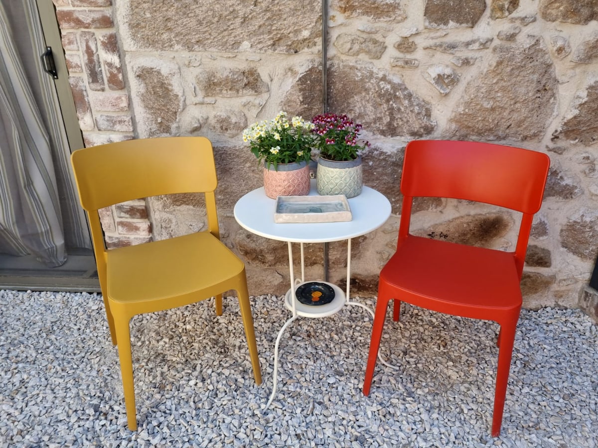 A small white table is positioned between two colorful chairs, one yellow and one red. Potted flowers in pastel-colored containers add a touch of nature, enhancing the outdoor seating area against a backdrop of rustic stone walls.
