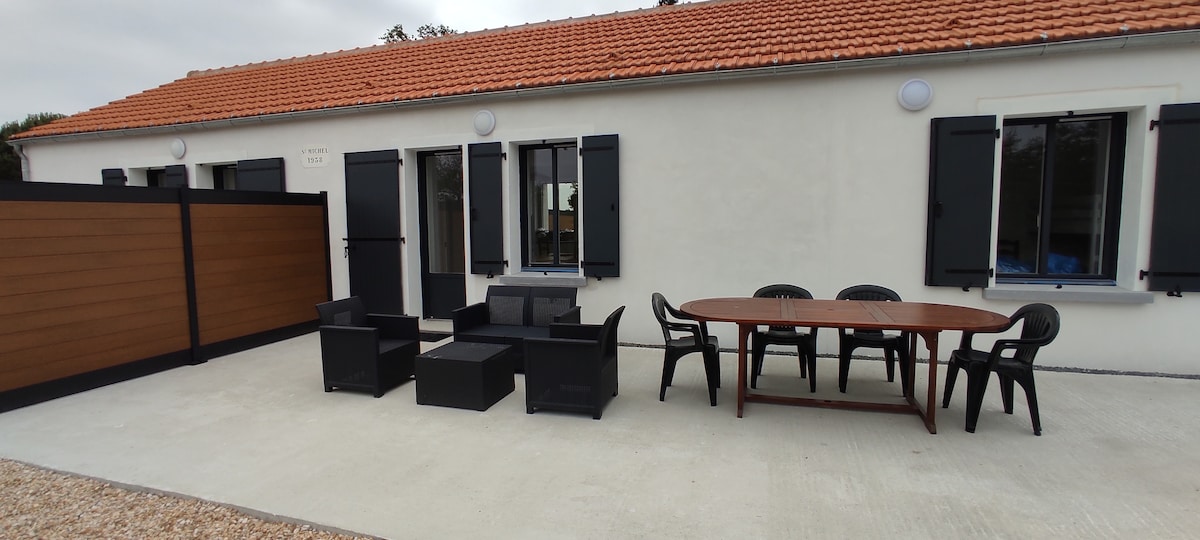 An outdoor terrace features a large table with several black chairs, accompanied by a lounge arrangement of dark seating. Windows with shutters and light-colored walls create a simple, functional space, and a shaded area is evident to provide comfort.