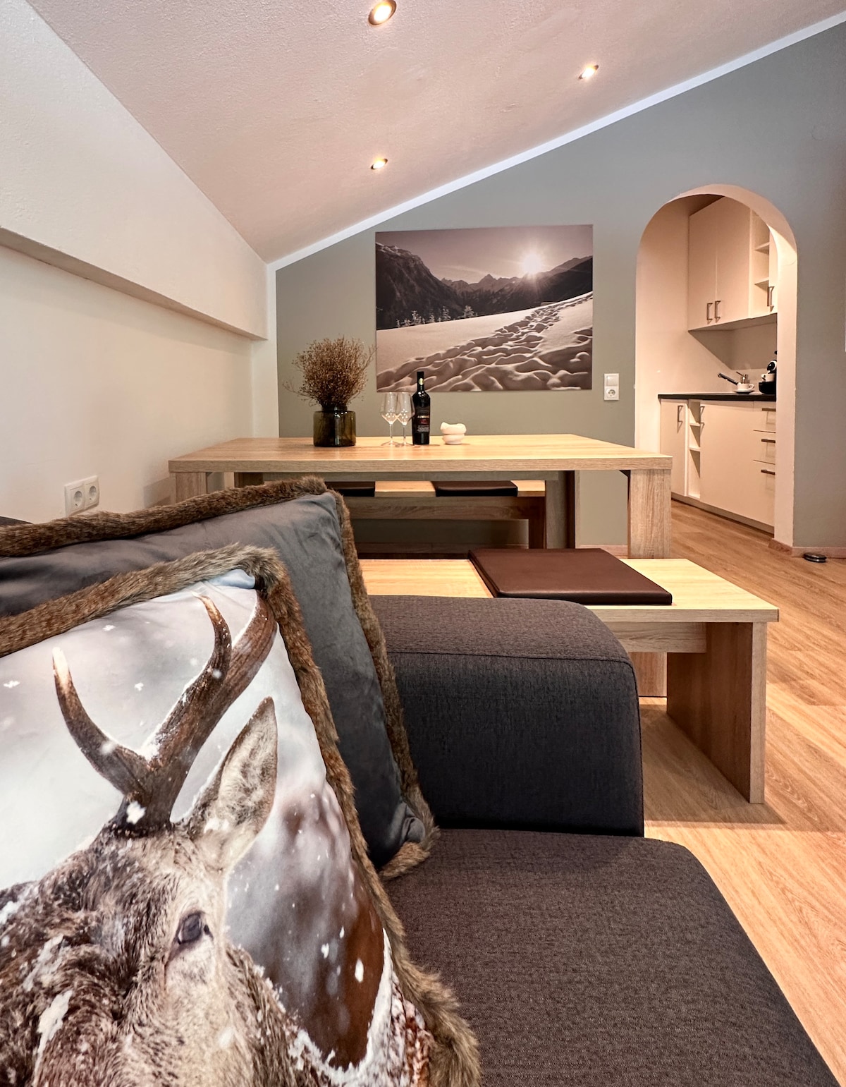 A cozy living area is highlighted by a dark gray sofa adorned with a decorative cushion featuring a deer. A wooden dining table is positioned nearby, complemented by a minimalist kitchen visible in the background. Natural light is present, enhancing the neutral color palette of the space.