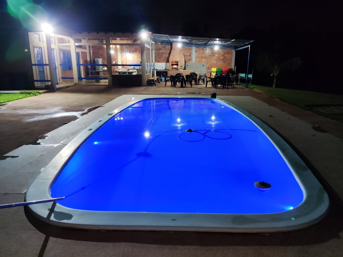 A well-lit swimming pool glows in blue, surrounded by a spacious patio. Tables and chairs are arranged nearby, and a covered area provides additional seating. The serene landscape is complemented by nearby greenery, creating a relaxing outdoor setting.