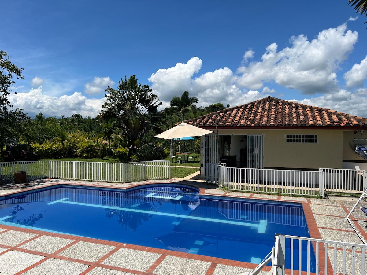 A serene outdoor space features a crystal-clear swimming pool bordered by a white fence. Lush greenery surrounds the area, and a shaded seating area is present under an umbrella near the pool. A charming villa with a tile roof stands in the background.