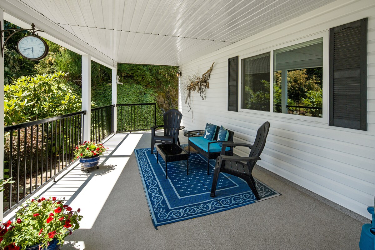A front porch area is furnished with two chairs and a small table, set against a backdrop of lush greenery. A decorative clock is mounted on the wall, and potted flowers add vibrant color to the space. Sunlight casts gentle shadows on the textured surface.