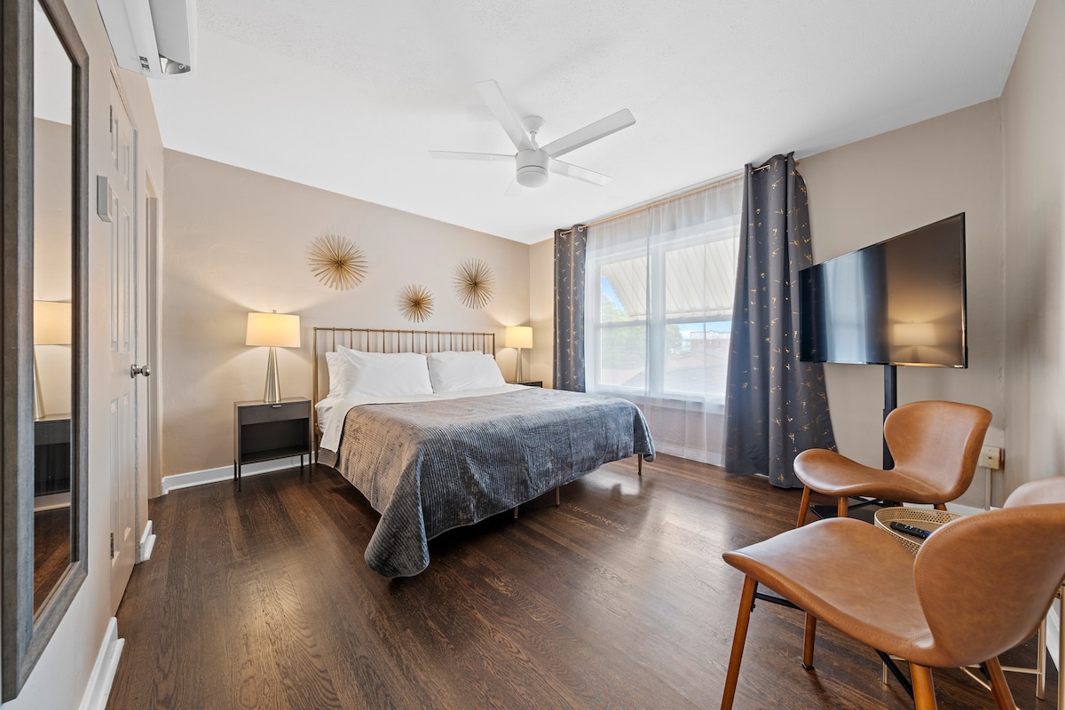 A spacious bedroom is showcased with a comfortable bed, accompanied by two bedside lamps illuminating the area. A large window allows natural light to enter, while a ceiling fan and flat-screen TV are also present. Two wooden chairs are positioned nearby for seating.