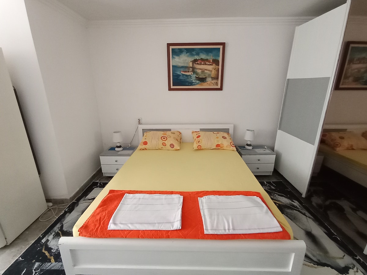 A spacious bedroom is presented with a light yellow bedspread and accent pillows featuring a floral pattern. Two bedside tables with lamps are positioned on either side of the bed. A decorative painting hangs above, and a mirrored wardrobe enhances the sense of space.