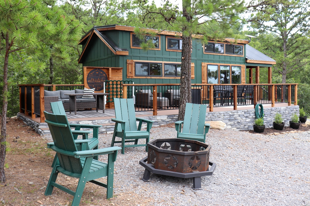 The modern tiny home is surrounded by trees, featuring large windows and a beautiful deck. A fire pit with metal grate is positioned in front, accompanied by four green Adirondack chairs. The setting provides a tranquil retreat atmosphere with natural landscaping.