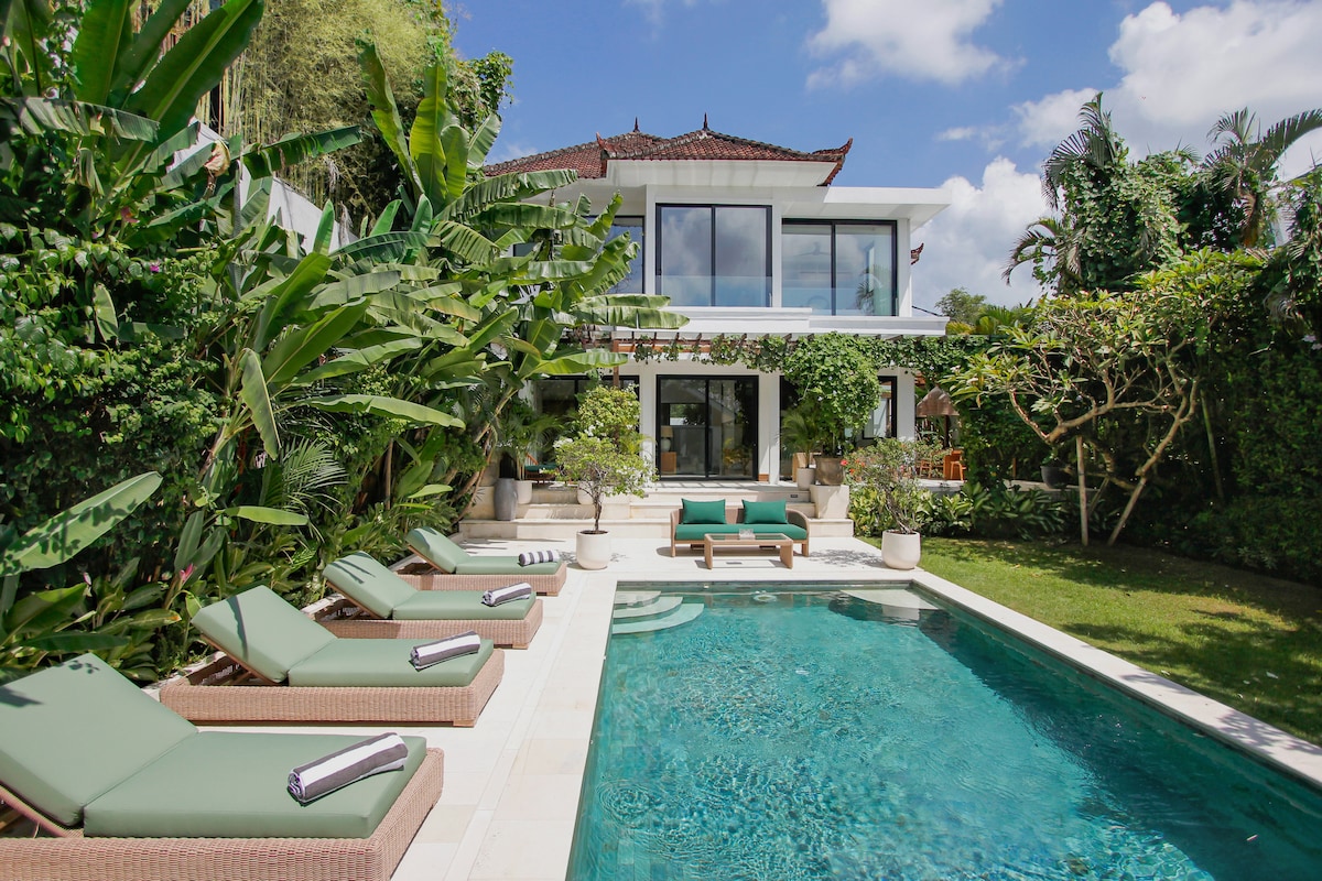 A private pool is bordered by lush tropical plants and green sunbeds. The villa structure showcases a mix of modern and traditional Balinese architecture. Large glass doors create a seamless transition between indoors and the outdoor lounging area, offering a sense of spaciousness.