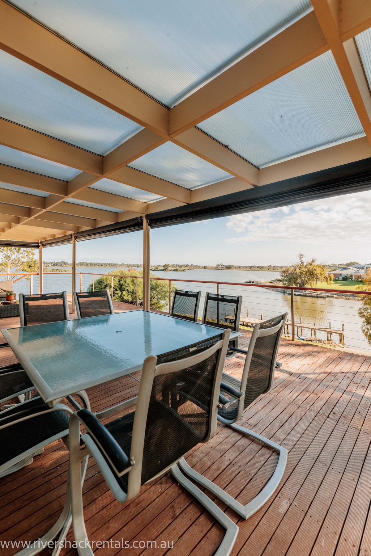 An outdoor dining area is presented on a spacious deck, featuring a large glass-top table surrounded by multiple chairs. Sunlight filters through a translucent roofing structure, illuminating the scenic views of the river and surrounding landscape.