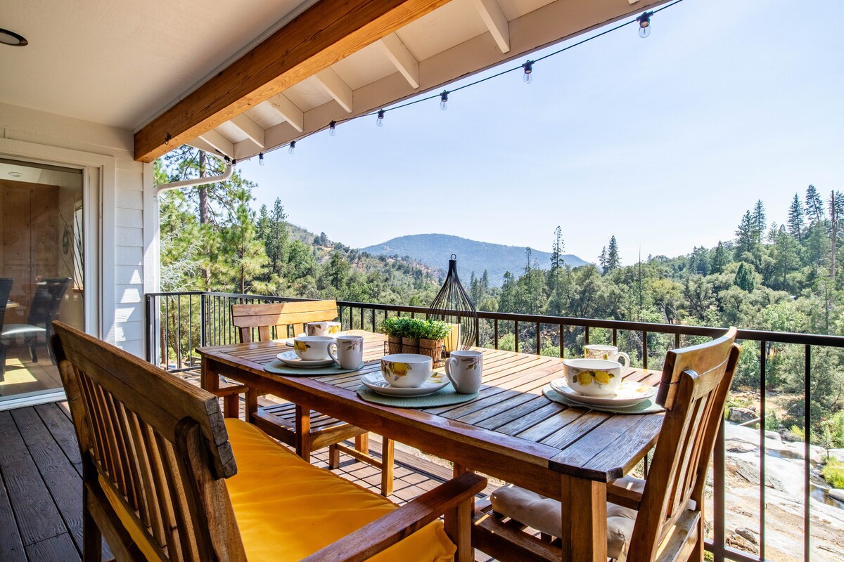 An outdoor dining area is showcased with a wooden table set for four, featuring decorative cups and a small potted plant in the center. The backdrop reveals scenic mountain views and lush greenery under a clear blue sky, enhanced by warm sunlight.