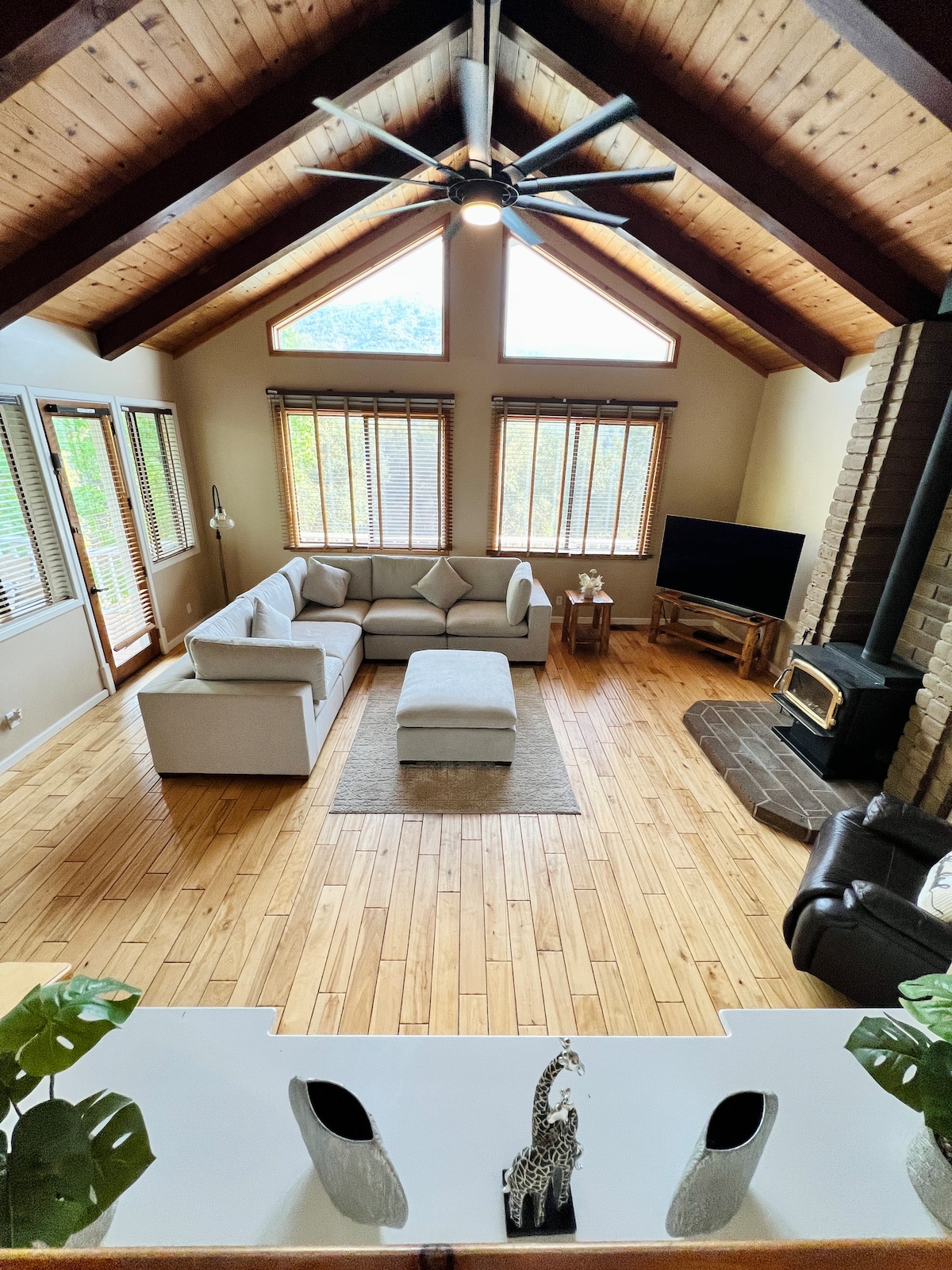 A spacious living area features high vaulted ceilings with wooden beams and a large window setup that allows natural light to fill the room. Comfortable seating is arranged around a central ottoman, with a wood stove and a TV situated in the corner.