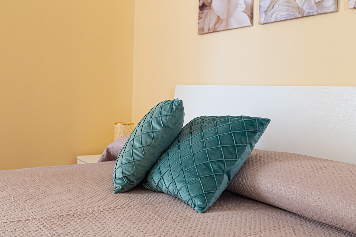 A cozy sleeping area is presented with a large, textured coverlet in a warm tone. Two plush, green cushions are positioned at the back of the spacious bed, complemented by light walls featuring subtle artwork above.