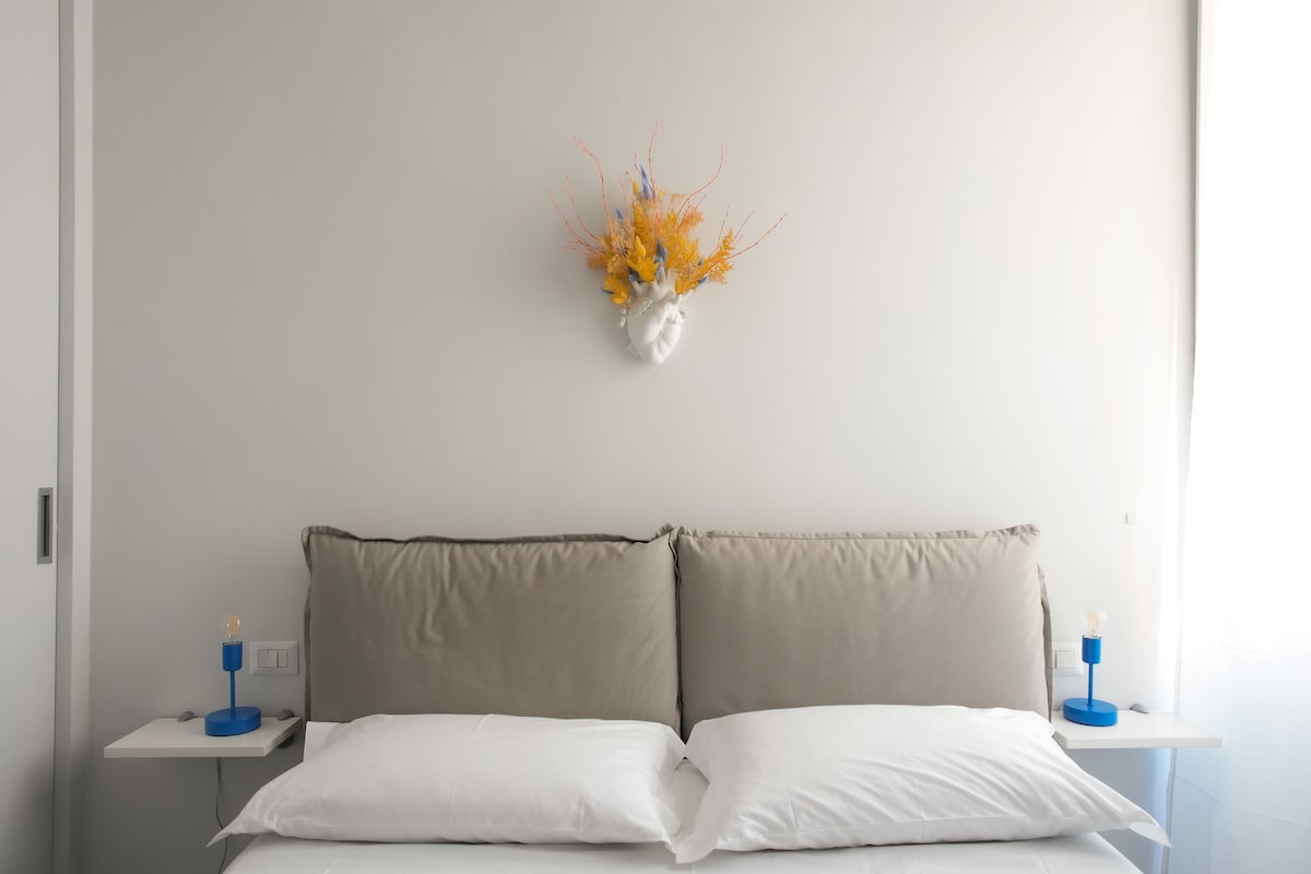 A neutral bedroom features a spacious bed with soft, light bedding, complemented by two bedside tables. Each table is adorned with colorful lamps. A floral wall decor piece in warm tones adds a cheerful touch above the headboard, while natural light filters softly through the window.