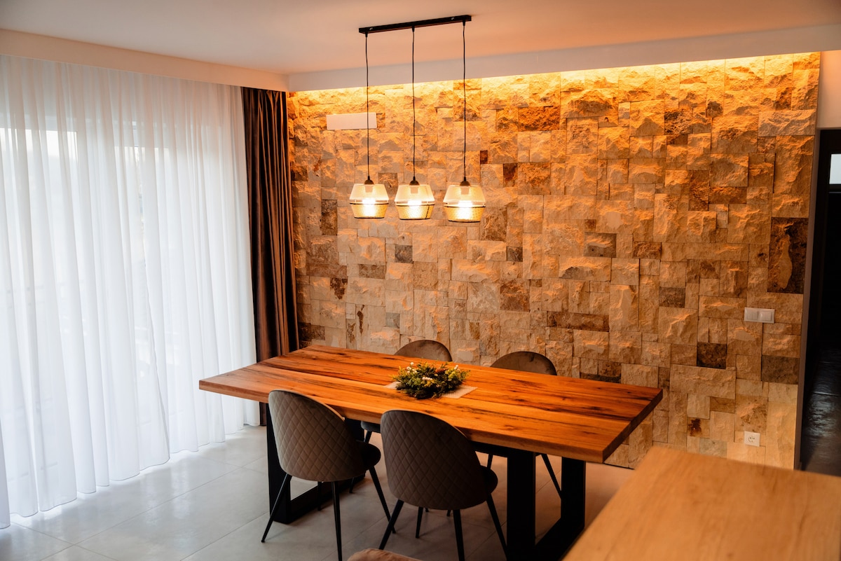 A dining area features a large wooden table surrounded by four upholstered chairs. Natural stone walls add texture, while warm lighting from three pendant lamps creates a cozy setting. Sheer curtains allow soft light to filter in, enhancing the serene atmosphere.