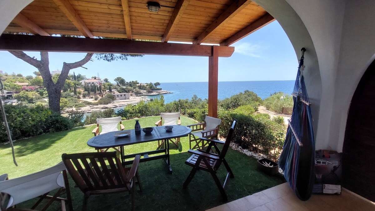 An outdoor patio area features a wooden table surrounded by chairs, overlooking the serene sea. Lush greenery frames the view, while gentle waves can be seen in the distance. A hammock is casually hung nearby, inviting relaxation in the coastal breeze.
