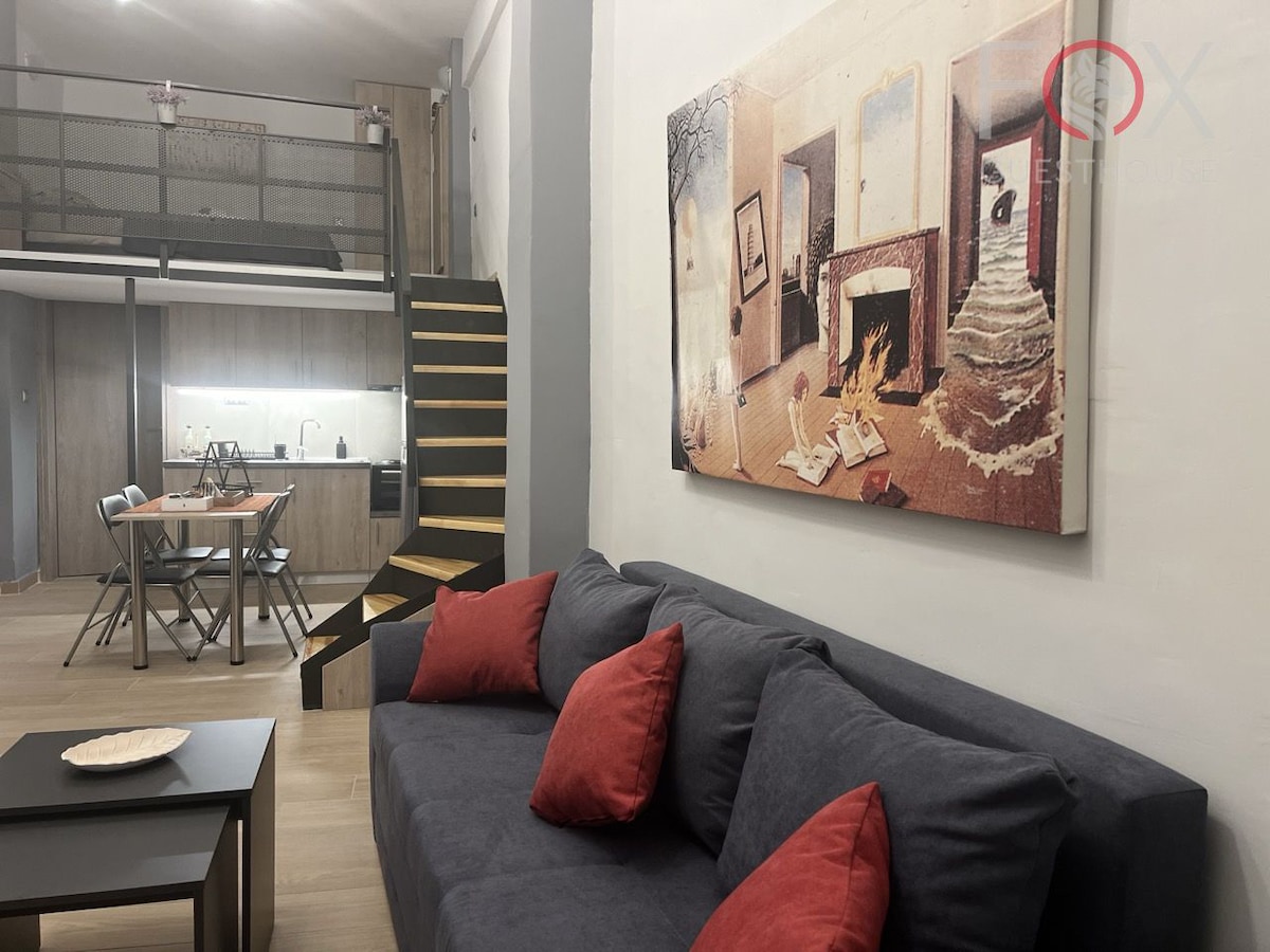 A modern living area features a comfortable dark sofa adorned with red cushions. A large wall art piece depicting a cozy interior scene adds visual interest. In the background, a dining table with chairs is positioned next to an open kitchen, and a staircase leads to an upper level.