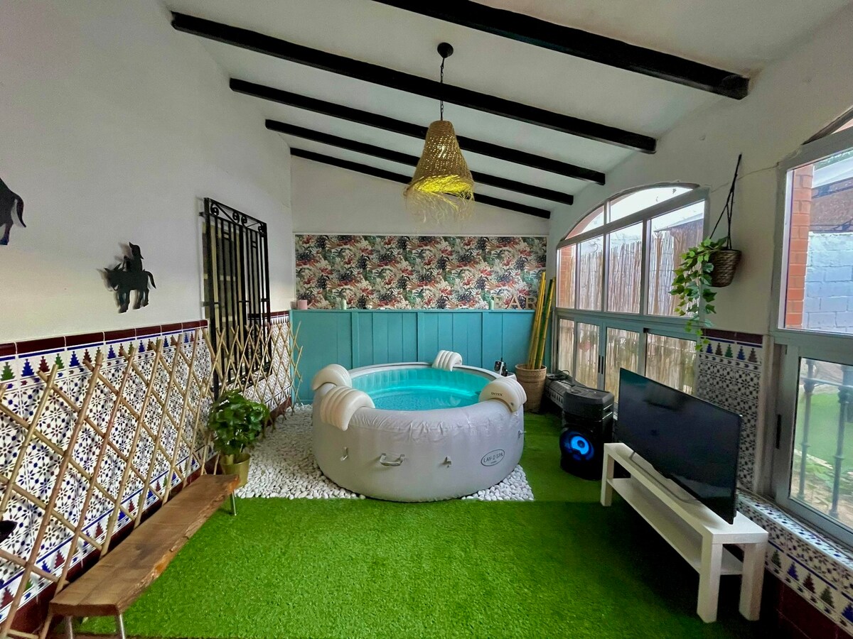 An indoor space is showcased, featuring an inflatable hot tub surrounded by decorative wall patterns and lush green artificial grass. A flat-screen TV is positioned nearby, and natural light floods the area through large windows, complemented by a woven pendant light hanging from the ceiling.