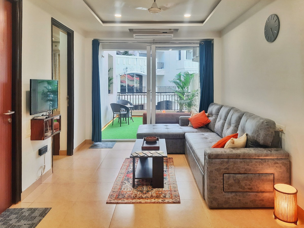 A spacious living room features a large, plush sofa paired with vibrant cushions and a coffee table. Natural light fills the space through sliding glass doors that open to a balcony with outdoor seating. A decorative rug adds warmth to the tiled floor.