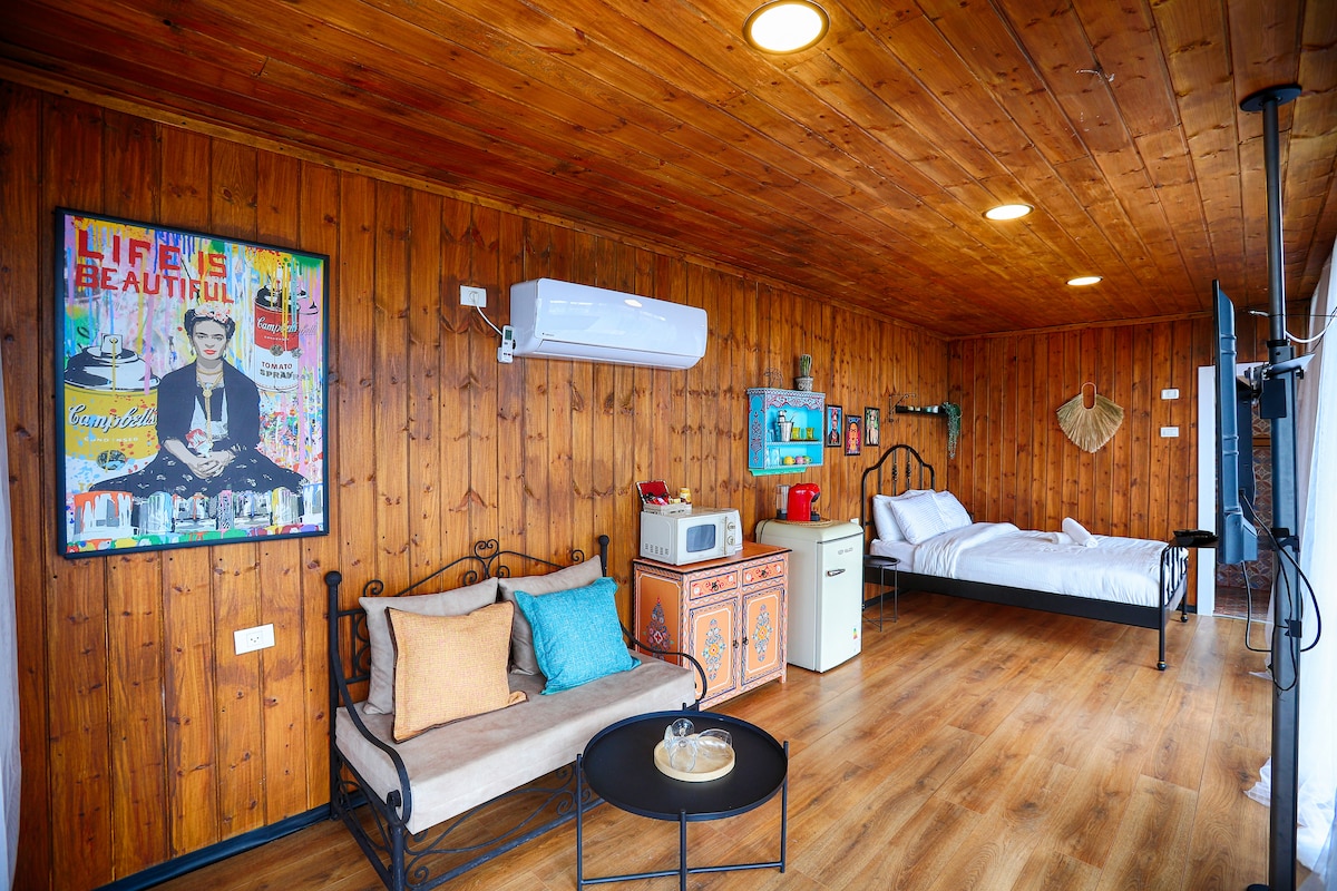 The studio suite features a wood-paneled interior with a spacious bed and a sitting area equipped with a small sofa. A microwave and mini-fridge are present, alongside a decorative vintage-style cabinet. Ambient lighting highlights the art on the walls.