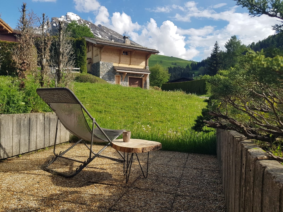 A terrace area is shown with a reclining chair positioned on a gravel surface. Lush green grass and sparse shrubbery create a natural setting, accompanied by a distant view of mountainous landscape and partly cloudy sky.