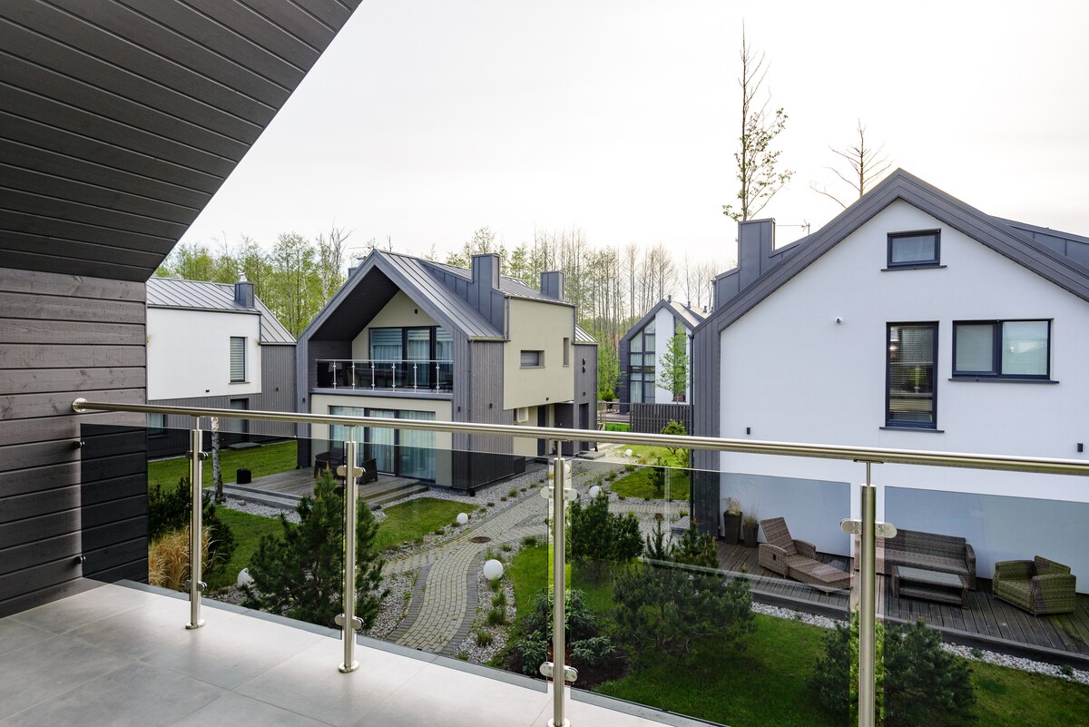 A balcony overlooks a landscaped area with modern houses and greenery. A pathway winds through the yard, bordered by various shrubs and plants, enhancing the serene environment. The architectural design features a mix of dark and light exteriors, creating visual contrast.