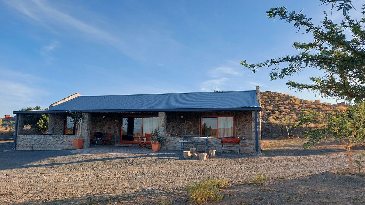 A stone cottage with a metal roof is showcased against a clear blue sky. Large windows reflect natural light, while planters with greenery add a touch of nature. The surrounding landscape features gentle hills, enhancing the serene atmosphere of the location.