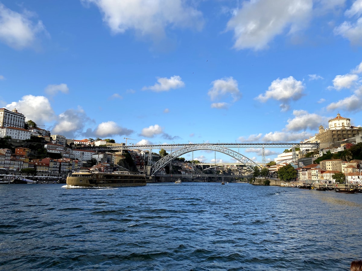 A panoramic view of the Douro River, framed by colorful buildings along the riverbank. A large bridge spans the river, connecting the two sides. The sky is partly cloudy, with blue patches peeking through. Water ripples gently in the foreground.