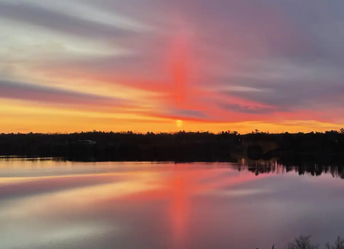A stunning sunrise casts vibrant hues of pink and orange across the tranquil surface of Boardman Lake. The reflections in the water enhance the serene landscape, with gently rolling silhouettes of trees and distant land visible along the horizon.