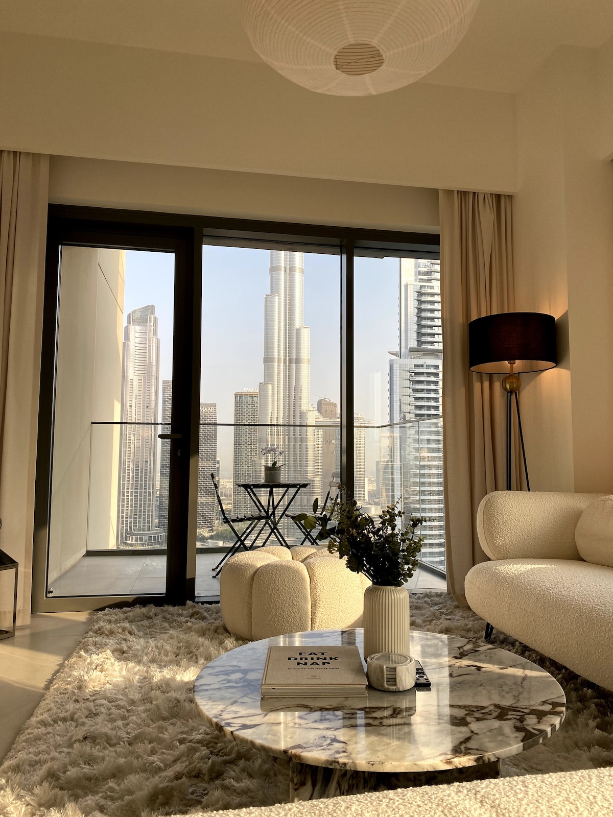 A modern living space features floor-to-ceiling glass doors that open to a private balcony. A round marble coffee table sits on a plush area rug, complemented by a small outdoor seating set. The iconic Burj Khalifa is prominently visible through the glass.
