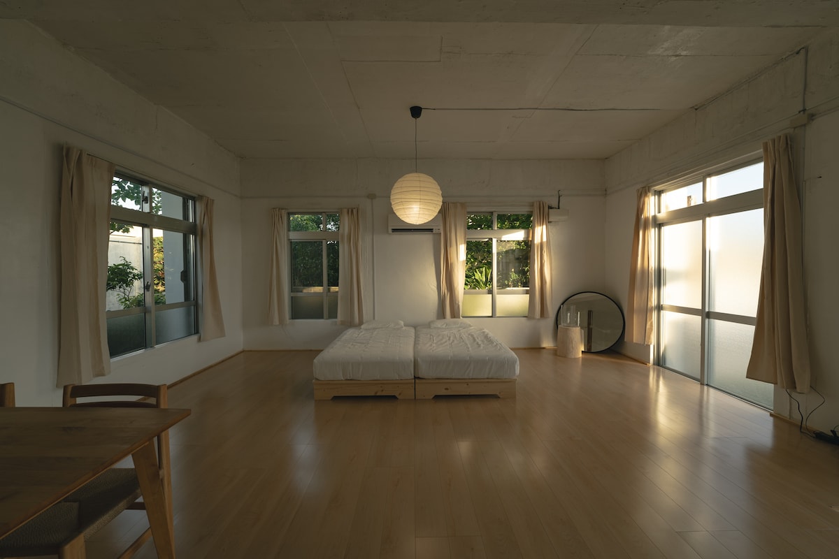 The interior space is characterized by wooden flooring and large windows allowing natural light to enter. Two beds are positioned in the center, surrounded by minimalistic decor. A dining table occupies one corner, and soft curtains frame the windows, enhancing the serene environment.