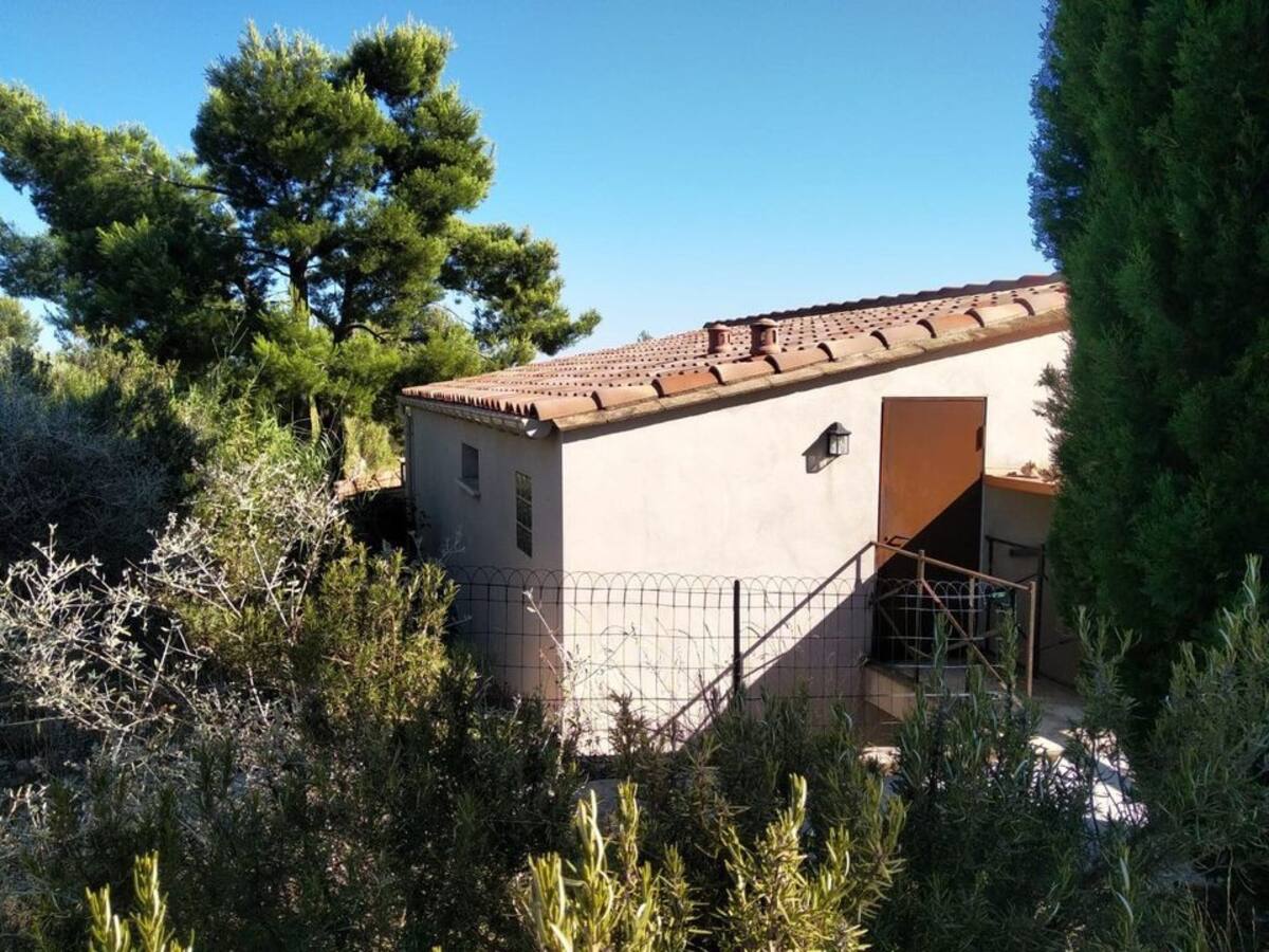 A rustic building is nestled among greenery, featuring a simple façade and a terracotta-tiled roof. An entrance door is visible, framed by lush shrubs and trees, creating a sense of privacy and seclusion in a natural setting.