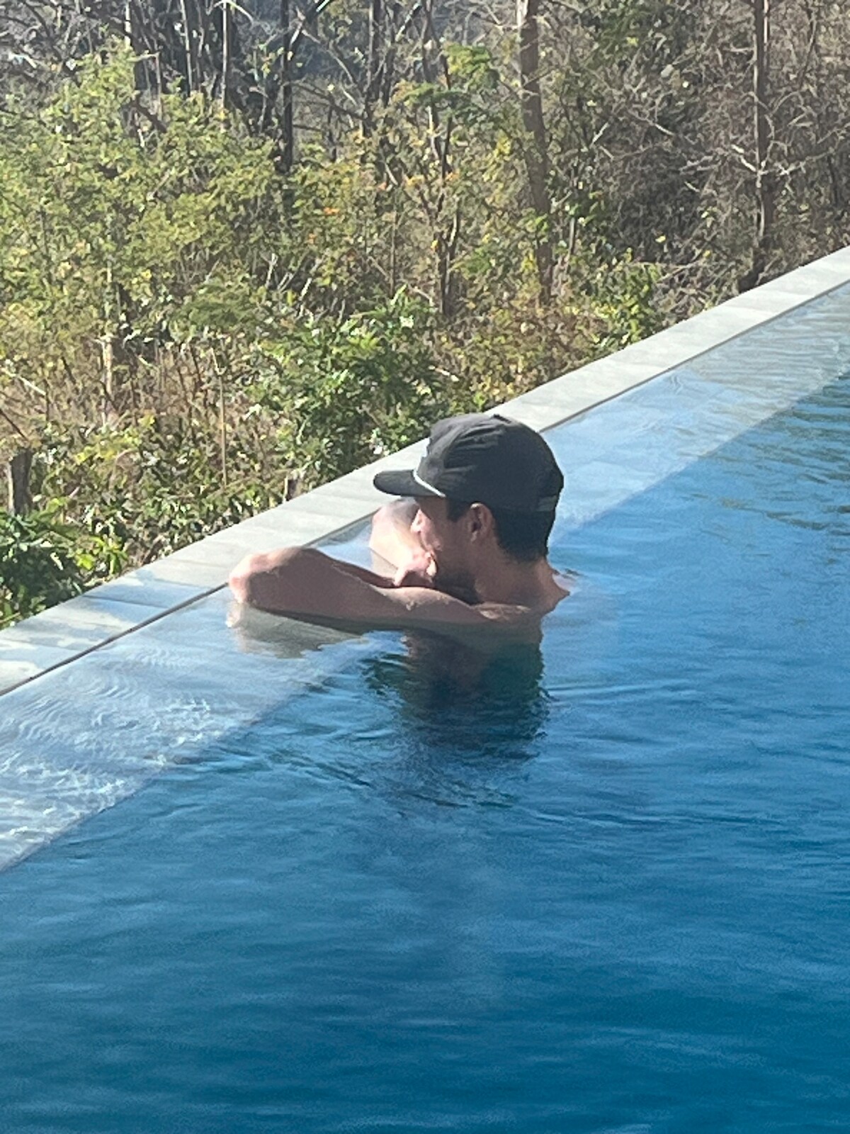 A person is seen relaxing in the edge of an infinity pool, surrounded by lush greenery. The water reflects the sky, creating a serene atmosphere. Towering trees and foliage in the background enhance the sense of privacy and tranquility.